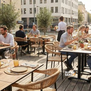 Super-Seat | HPL Hospitality Tischplatten Castle Oak 80x80cm Hölzerne eichenfarbene Tische mit Stahlrahmen und Stühlen, umgeben von Pflanzen und Leuchten. | Catering Möbel