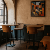 Super-Seat | Eine kleine Café-Ecke mit zwei Holztischen, mehreren Barhockern im New Yorker Design, Mokka-Holzvelours und Holzstühlen, einer Glasflasche und einem Glas auf einem Tisch, einem großen Bogenfenster auf der linken Seite und einem bunten abstrakten Gemälde an der Wand. | Holztische, Mokka-Velours-Barhocker mit Holzbeinen, Holzstühle; warme Töne und Glasdekor. | Catering Möbel