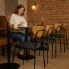 Super-Seat | Eine Frau sitzt allein an einem runden Tisch in einem Café | Schwarze und hölzerne skandinavische Stühle, runde Holztische, Trockenblumen in Vasen. | Catering Möbel