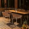 Super-Seat | Ein hölzerner Esstisch für draußen mit sechs Ibiza wit (terras) stapelstoelen met kussen | Lichtbruine houten tafel, witte kunststof stapelstoelen met grijze kussens, glazen fles, planten. | Horecameubilair