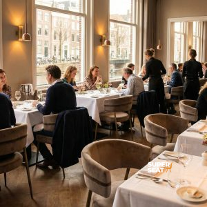 Super-Seat | Mounira Design Samtige Mokka-Sessel mit goldenen Details, weiße Tische und Blumenarrangements in einem hellen Restaurant am Wasser. | Catering Möbel